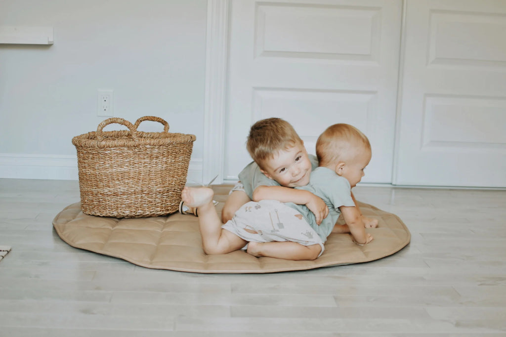 Two children sitting on a wooden mat with a wicker basket in the background.