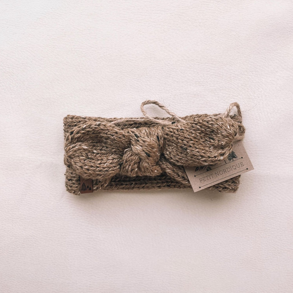 White background with Handknit Knot Bow Headband by Petit Nordique in Ochre Tweed. Headband has a knot bow at the front, with a small leather tag in an ochre tweed yarn.