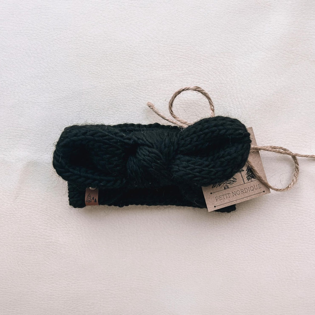 White background with Handknit Knot Bow Headband by Petit Nordique in Black. Headband has a knot in the front, with a small leather tag, in a black yarn.