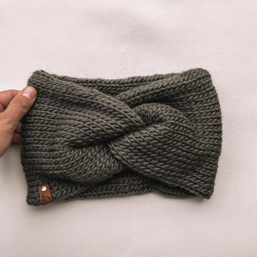 White background with a hand holding the Handknit Twist Headband by Petit Nordique in Olive. Headband has a twist at the front with a small leather tag, in an olive yarn.