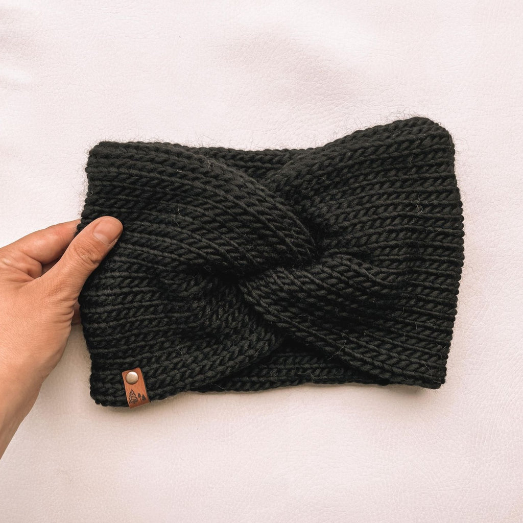 White background with a hand holding the Handknit Twist Headband by Petit Nordique in Black. Headband has a twist at the front, with a small leather tag, in black.