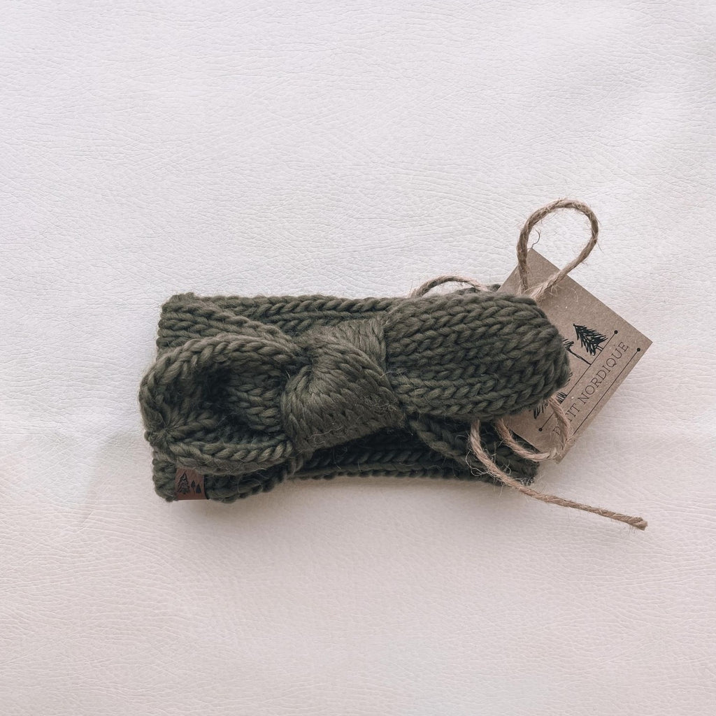 White background with Handknit Knot Bow Headband by Petit Nordique in Olive. Headband has a knot bow in the front, and a small leather tag in an olive yarn.
