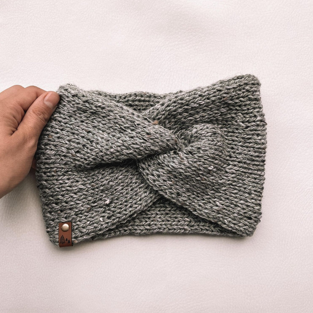 White background with a hand holding the Handknit Twist Headband by Petit Nordique in Grey Tweed. Headband has a twist at the front, with a small leather tag in a grey tweed yarn.